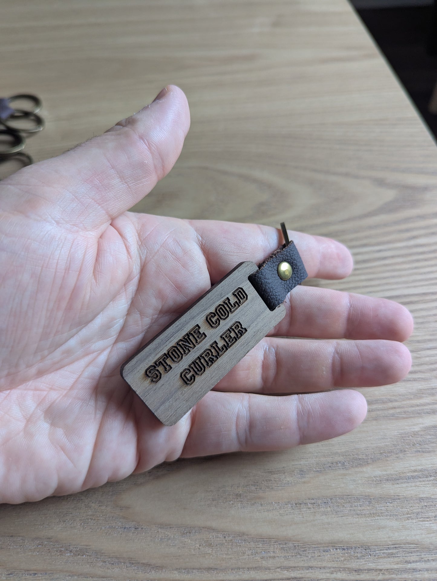 Hand holding a keychain with 'STONE COLD CURLER' engraved on a wooden surface